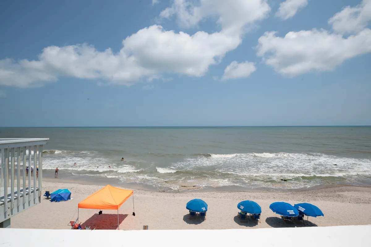 A scenic view of the beach with colorful umbrellas, an orange tent, rolling waves, and stretches of sand, evoking a sense of tranquility and the opportunity to observe nature and unwind by the ocean.