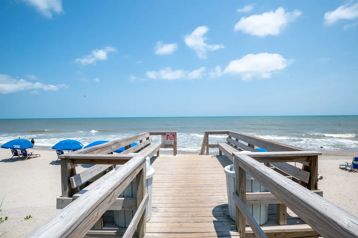 A tranquil view looking down a wooden walkway lined with beach umbrellas toward the ocean. The scene captures families sunbathing, swimming, and children building sandcastles along the shoreline, set against a backdrop of blue water and skies.