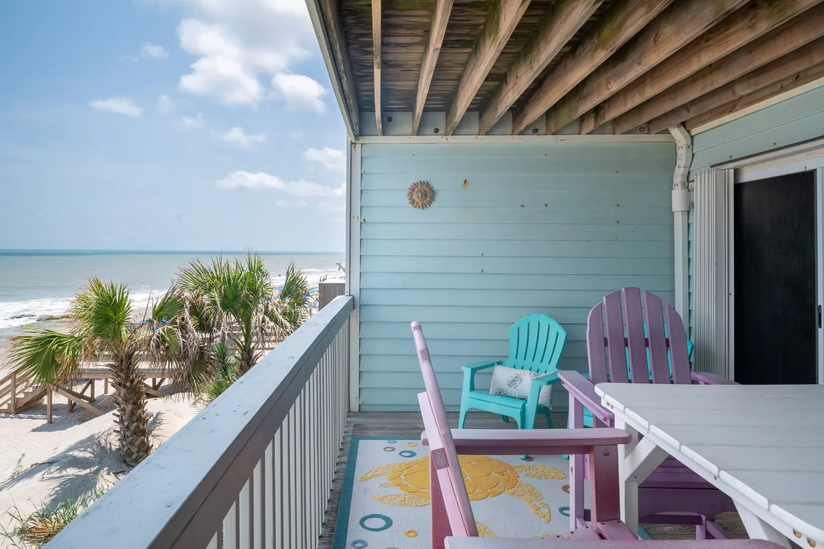 A breezy outdoor dining deck featuring a table surrounded by colorful chairs, overlooking beautiful palm trees and a panoramic view of the beach. The scene is tranquil yet vibrant, suggesting seamless indoor-outdoor living and al fresco dining options.