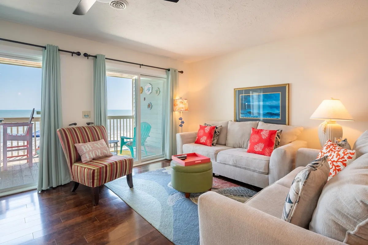 A comfortable living space with a sofa featuring red pillows, a striped armchair, a vibrant rug, and floor-to-ceiling windows offering an ocean view. The atmosphere is cheerful and perfect for socializing or unwinding while enjoying natural light.