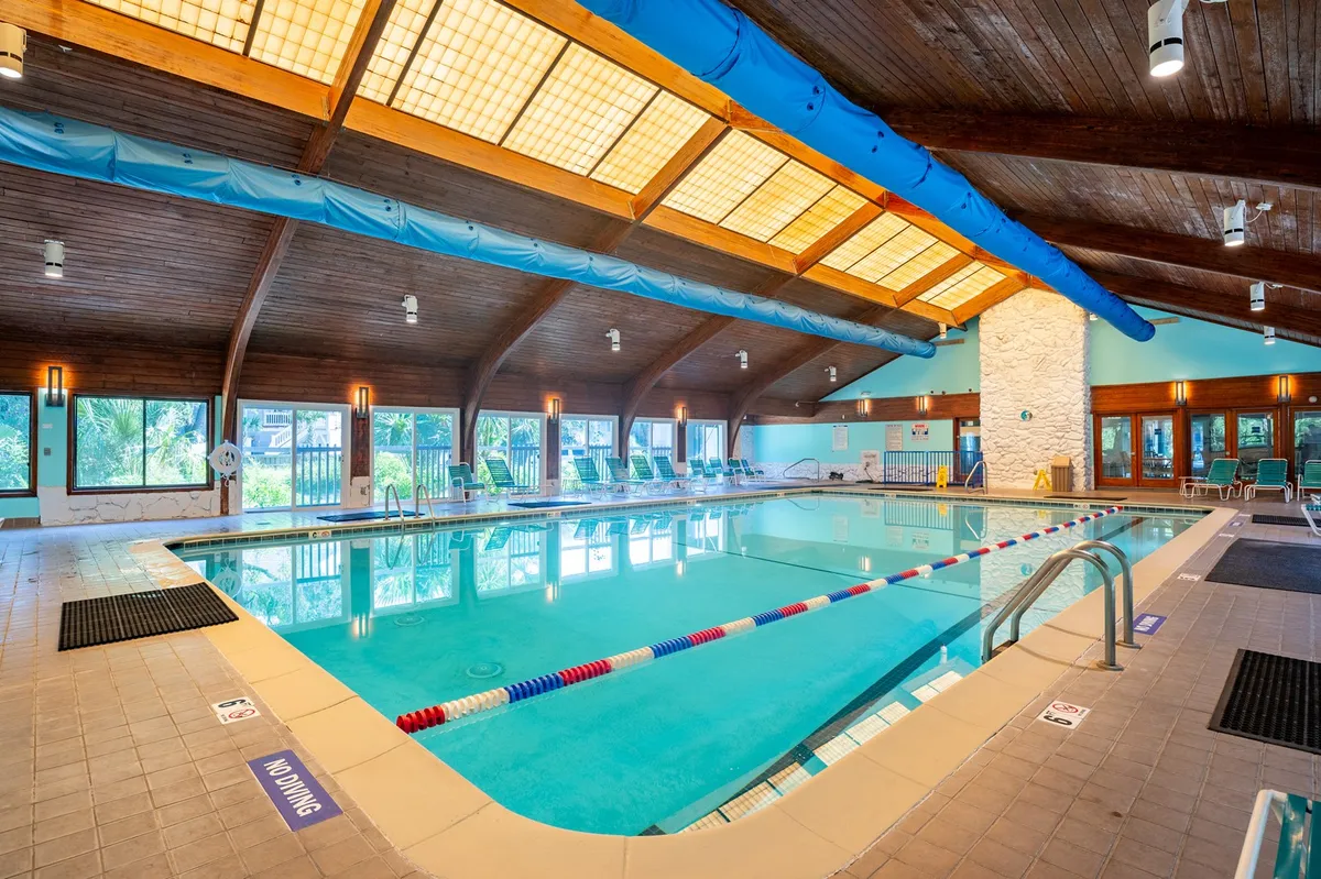 An indoor swimming pool with a wooden ceiling, sunlight streaming through large windows, and a peaceful ambiance. The space is inviting for both relaxation and exercise, and the design hints at a high-quality resort environment.
