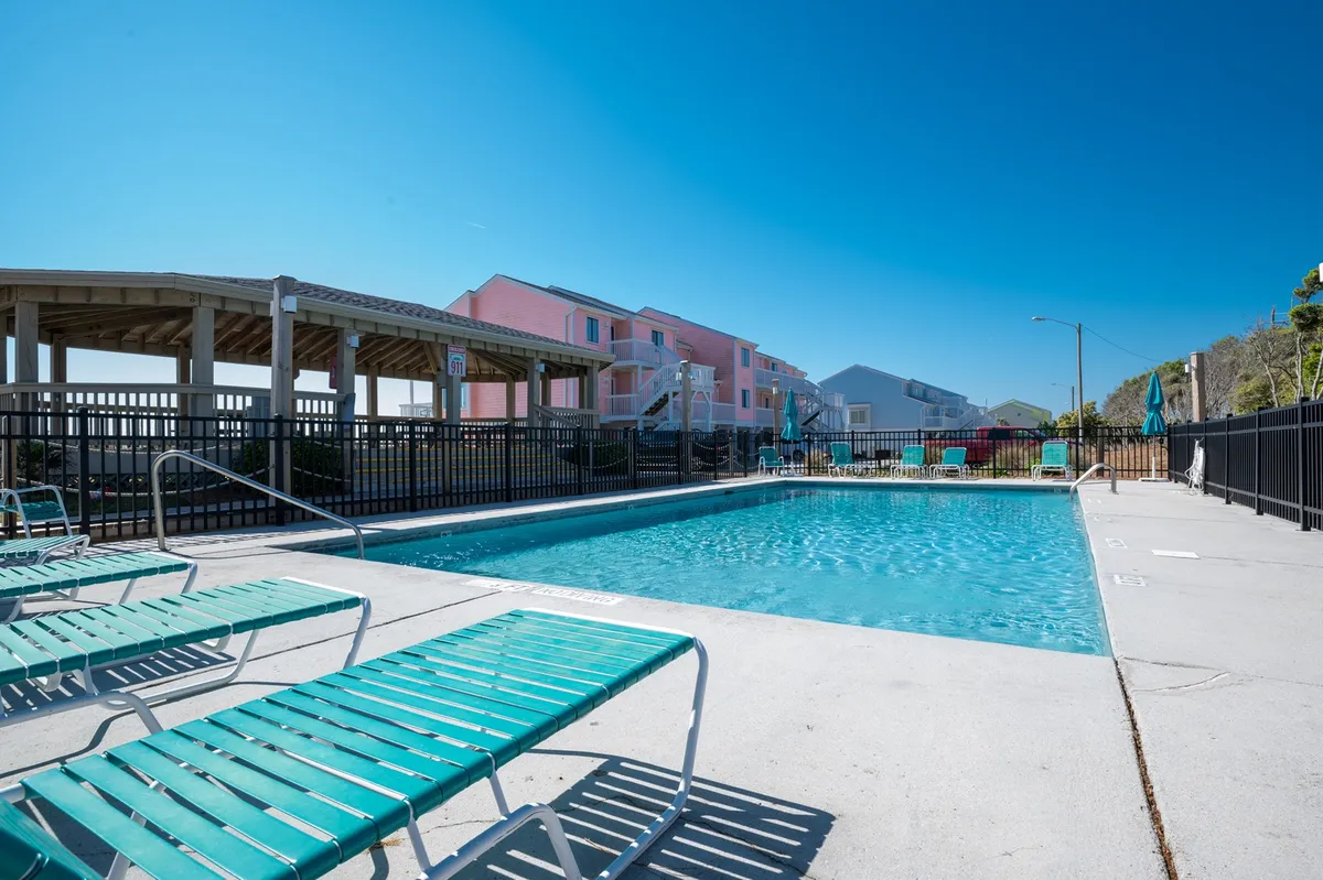 A bright, inviting exterior of one of the resort's swimming pools surrounded by lounge chairs and adjacent buildings, with families swimming, sunbathing, and relaxing poolside. The relaxed atmosphere and open layout encourage community and family gathering.
