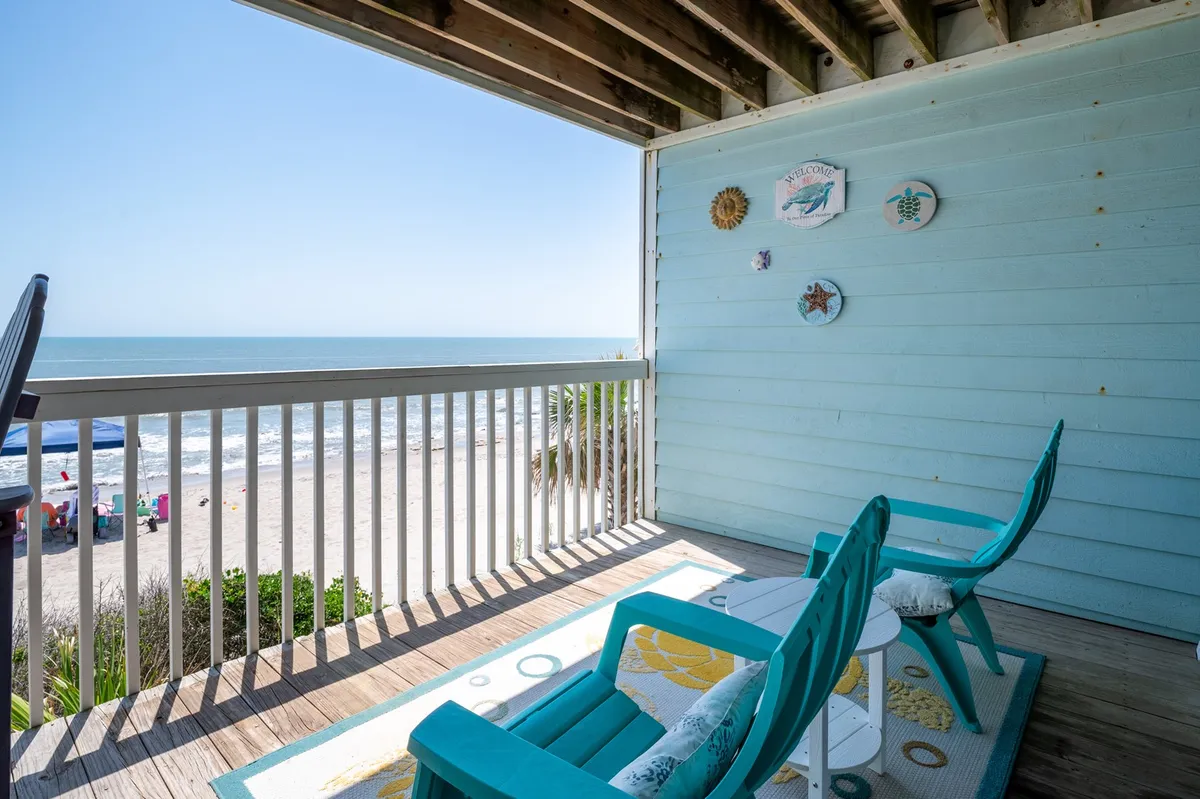 A tranquil exterior view featuring turquoise chairs on a deck, decorative wall plates, and a sweeping beach panorama beyond. The setting suggests a peaceful space perfect for sipping morning coffee or relaxing at sunset, with direct connection to the ocean breeze.
