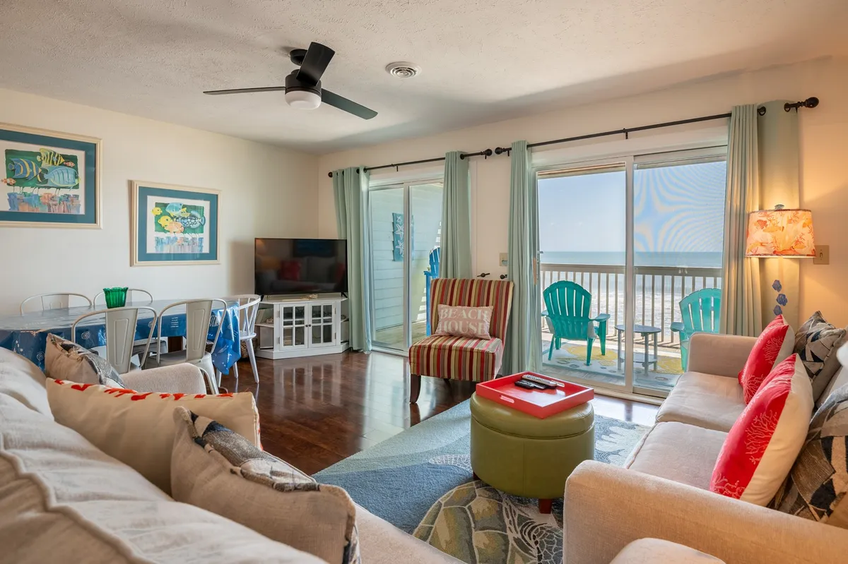 A cheerful, casual living space with a sofa adorned with colorful cushions, a dining table set for gathering, and a view of the ocean through sliding doors. A striped chair and playful 'BEACH HOUSE' sign add to the vacation vibe. The scene feels ideal for relaxing, watching TV, or sharing meals as a group.