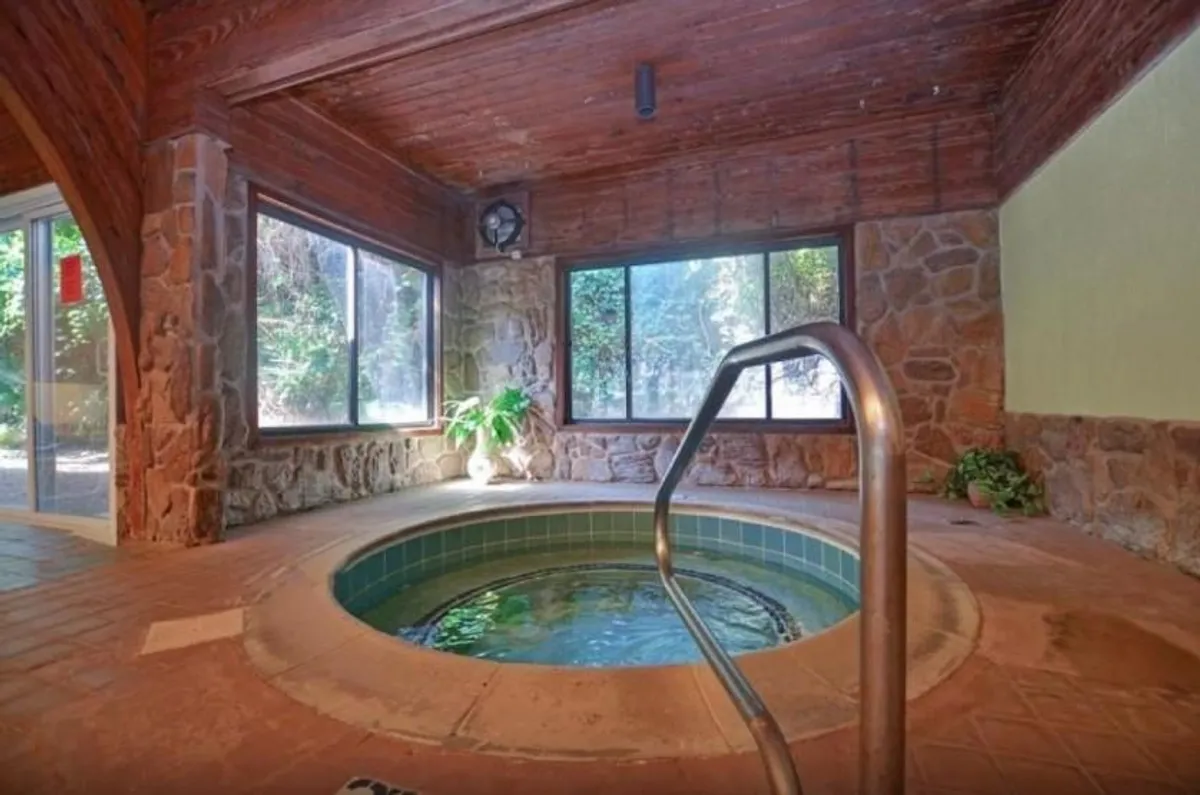 An elegant indoor spa room highlighted by a bubbling hot tub, natural stone walls, large windows, and a wooden ceiling. The space feels secluded, luxurious, and designed for adult relaxation and wellness.