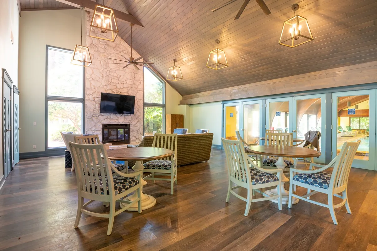 A spacious open-concept living and dining area, featuring large windows that bathe the space in natural light. There's a cozy stone fireplace, hanging light fixtures, and comfortable dining tables and chairs. The overall mood is welcoming and ideal for social gatherings and family events.