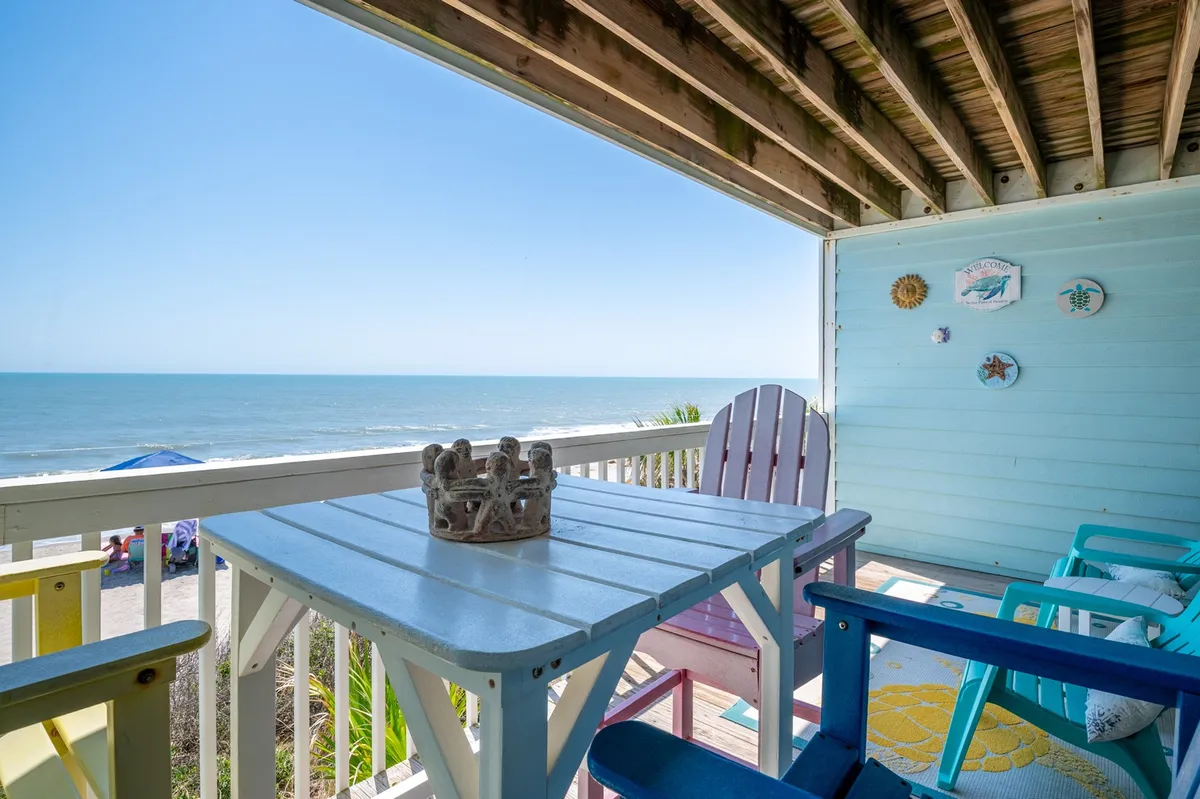 The image features a tranquil oceanfront balcony with colorful chairs and a table adorned with a crown decoration, all overlooking the sparkling Atlantic. The inviting seating is perfect for sunrise coffee, relaxed afternoons reading, or gathering with friends to watch the waves roll in.