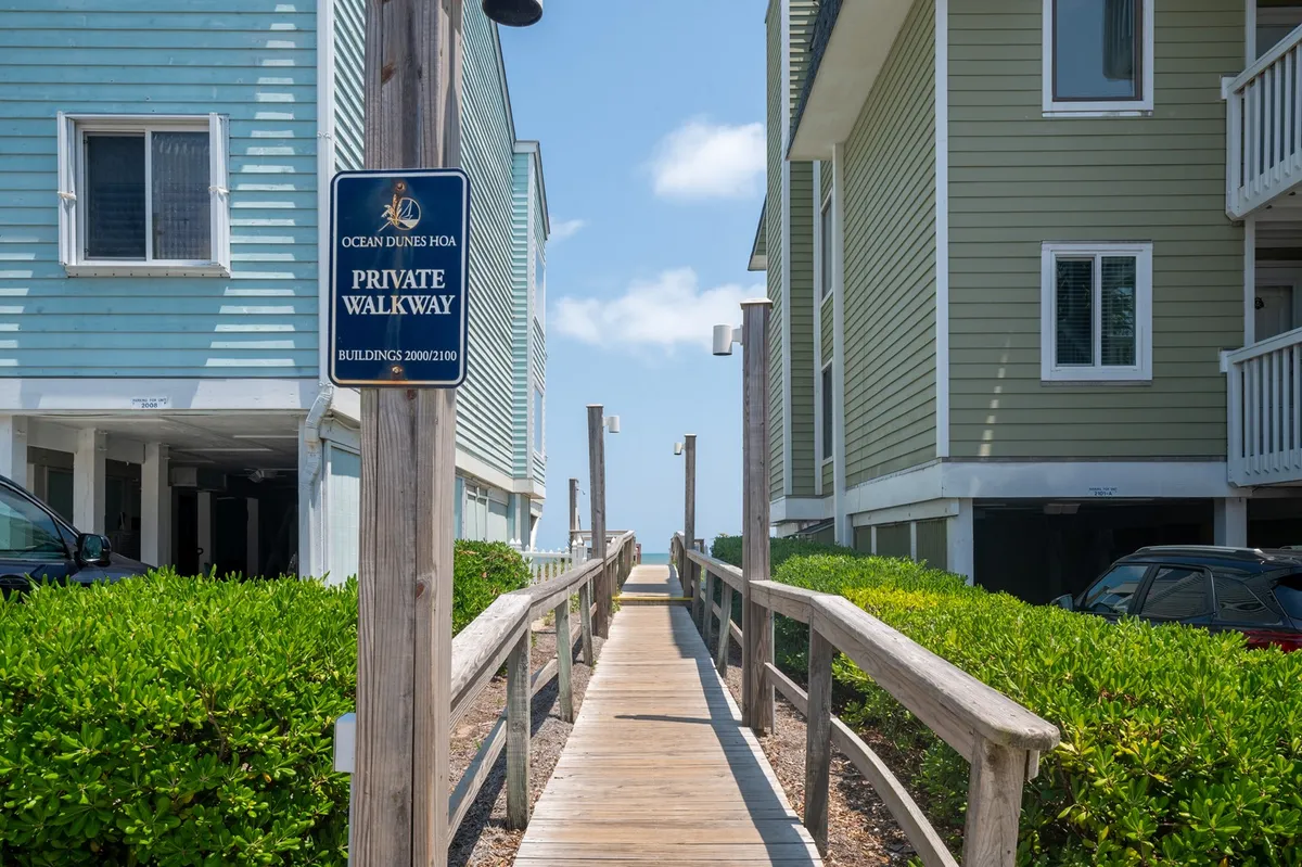 A tranquil wooden walkway flanked by lush greenery, leading toward the ocean, with a 'private walkway' sign and glimpses of the natural landscape and adjacent buildings.